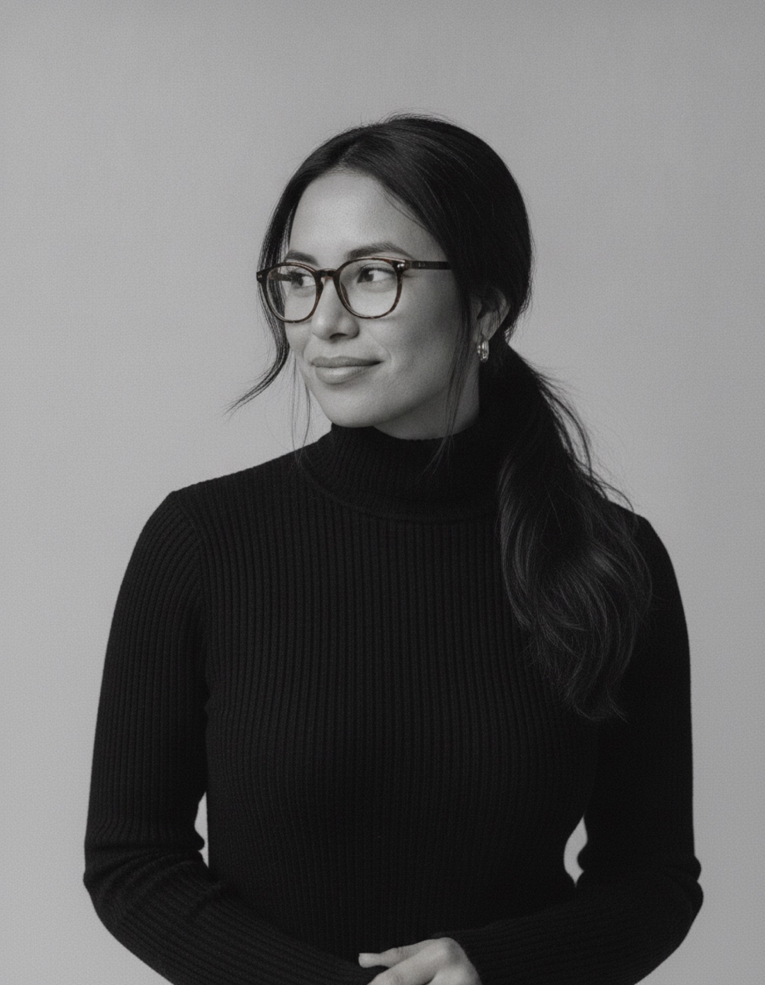 Black and white portrait of Maya Reyes in a black turtleneck and glasses, looking to the side with a small smile